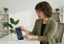 A professional tech reviewer in an olive green blazer holds the Samsung Galaxy S26 Ultra in Titanium Gray, smiling and looking at the screen for an in-depth review on Numerello.com. Modern home office background with bookshelves.